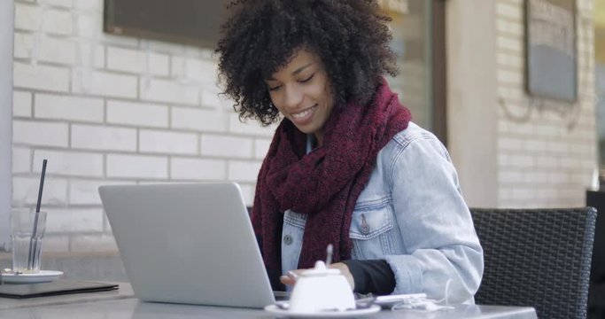 Attractive African woman in casual clothing laughing while sitting at table with laptop in cafe.