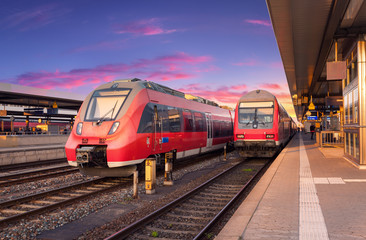 Naklejka premium Szybkie czerwone pociągi podmiejskie na stacji kolejowej i kolorowe niebo o zachodzie słońca w Europie. Krajobraz przemysłowy z pięknym peronem kolejowym i nowoczesnymi pociągami latem. Popędzać. Turystyka kolejowa