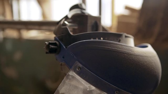 Close up footage in slowmotion of blue safety helmet with visor and ear pieces hanging on machine in joinery covered with wooden sawdust