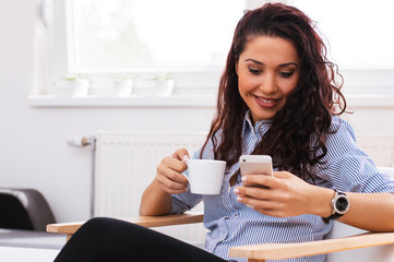 Portrait of young beautiful casual woman holding smartphone, looking at screen and holding cup of coffee