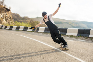 Longboarder on longboard in overalls helmet and gloves performs a stand-up slide at speed while on...