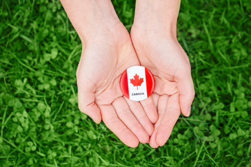 Macro closeup shot of hands palms holding round badge with red white canadian flag maple leaf on green grass forest nature background outside Canada Day celebration
