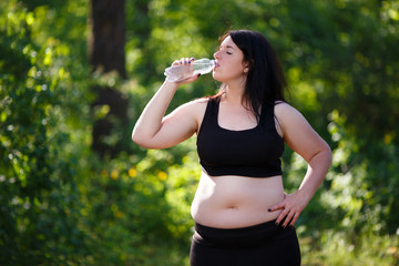 Young thirsty overweight woman drinking water after jogging in the park