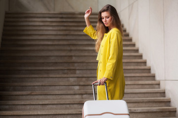 Young woman dressed in yellow suit holding suitcase and looking back at it, standing front of stairs