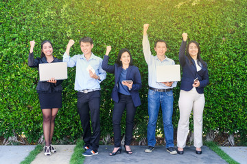Group of business people arm up and holding lab-top computer, tablet, smart phone standing in front of green tree leaves background, Business triumph, teamwork concept