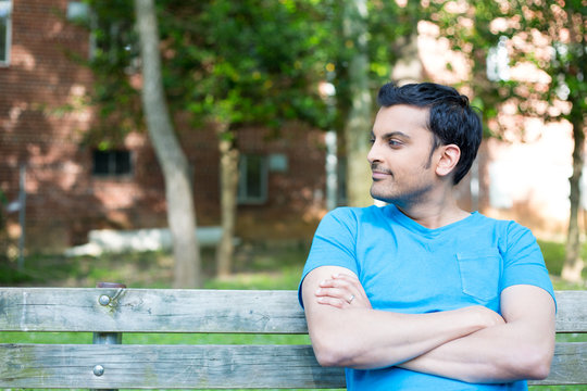 Closeup portrait, happy smiling, regular young man in blue shirt sitting on wooden bench, relaxed looking to side, isolate background trees, woods.