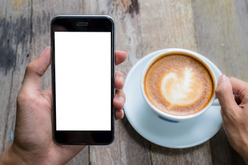 A man hand holding smart phone with latte hot, coffee cup on wood desk in coffee shop. Smartphone with blank screen and can be add your texts or others on smart phone.