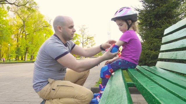 Father helps his daughter to wear a helmet and protective gear, for roller skating in the park.