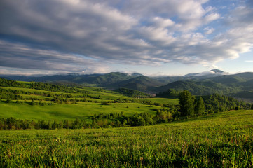 Valley among the green hills of the foothills with vivid bright meadows and groves of trees on the background of rural settlement and mountain ranges under the sunset sky.  Altai , Siberia, Russia.