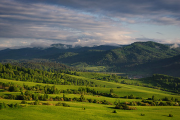 Fototapeta premium Valley among the green hills of the foothills with vivid meadows and groves of trees on the background of rural settlement and mountain ranges under the sunset sky. Altai, Siberia, Russia.