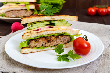 Multilayered sandwiches with a juicy cutlet, cheese, radish, cucumber, lettuce, arugula cutting in half on a plate on a dark wooden background. Close up