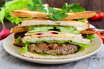 Multilayered sandwiches with a juicy cutlet, cheese, radish, cucumber, lettuce, arugula cutting in half on a plate on a dark wooden background. Close up