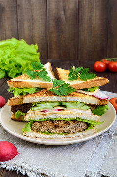 Multilayered sandwiches with a juicy cutlet, cheese, radish, cucumber, lettuce, arugula cutting in half on a plate on a dark wooden background. Vertical view