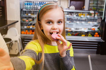 A little girl stands near a showcase with macaroon and makes selfie