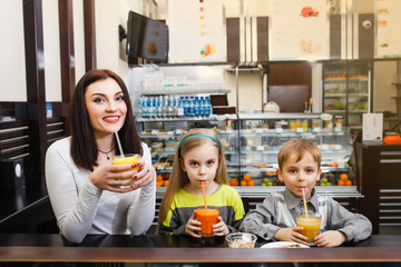 young Mom with her chidren is sitting in a cafe and drinking juice