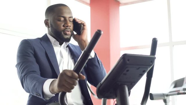 Serious male in suit exercising on stationary bike and talking on cellphone