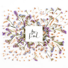 Words "Be Kind" written in calligraphic style on paper with blue and purple dried flowers on white background. Flat lay, top view