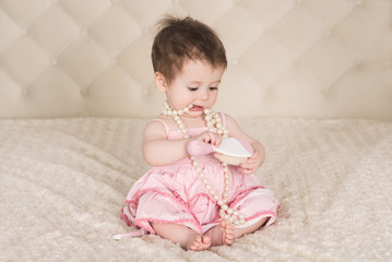 cute baby girl in pink dress, with pearl beads and comb in her hands sitting on the bed and playing, indoors