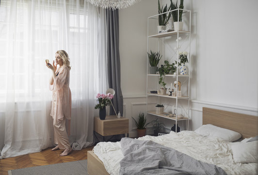 Pretty blonde Caucasian woman standing in her bedroom and putting make up on her face.
