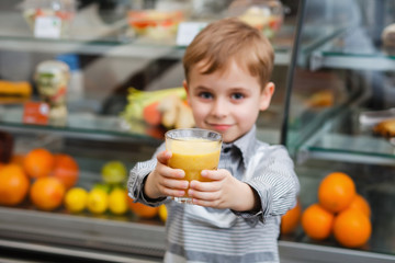 A little boy stands near a showcase with glass of juice in a cafe