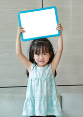 Cute little child girl holding empty white blackboard standing on fabric sofa in library room. Education concept.