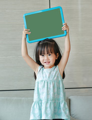 Fototapeta na wymiar Cute little child girl holding empty green blackboard standing on fabric sofa in library room. Education concept.