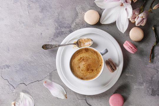 White cup of black coffee, served on white saucer with macaroons biscuits, spoon and magnolia flower blossom branch over gray texture background. Flat lay, space