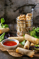 Fried spring rolls with red pepper sauce, served on paper and in fry basket with fresh green salad over old metal texture background. Top view. Asian food