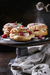 Homemade choux pastry cake Paris Brest with raspberries, almond and rosemary, served on black wooden serving board on cake stand over dark texture background. Sprinkling sugar powder. French dessert