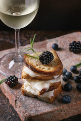 Grilled sandwich with melted goat cheese, blackberry, blueberry, rosemary and honey, served on terracotta board with glass of cold white wine over dark background. Summer appetizer.