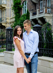 Fototapeta premium Young couple holding hands and walking through pathway on the street. Girl dressed in pink dress and man in blue shirt. Boy looking at the girl