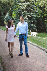 Fototapeta na wymiar Couple holding hands while walking in park. Girl dressed in pink dress and man in blue shirt and jeans.