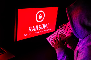 Computer screen with ransomware attack alert in red and a hacker man keying on keyboard in a dark room, ideal for online security failure and digital crime, long exposure selective focus