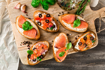 Italian bruschetta set. Varied sandwiches with cheese and red fish, tomatoes, fresh basil, spices, rosemary, garlic onion, chili pepper. Bottle with olive oil, rustic wooden board over dark background