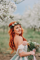 Young red-haired woman in a luxurious dress is standing in a blooming garden, close-up