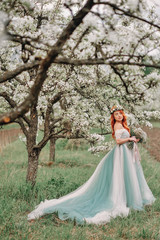 Young red-haired woman in a luxurious dress is standing in a blooming garden