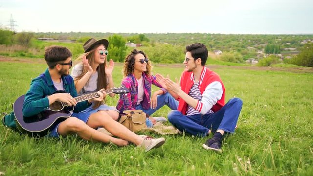 Group of stylish cheerful hipsters with guitar while sitting on a grass in the park. Summer leisure concept. Wide shot