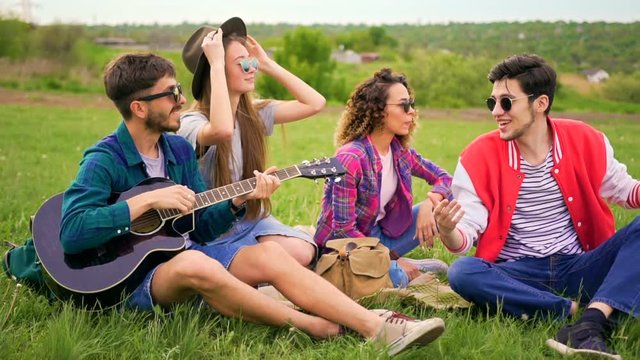 Group of stylish cheerful hipsters with guitar while sitting on a grass in the park. Summer leisure concept. Wide shot