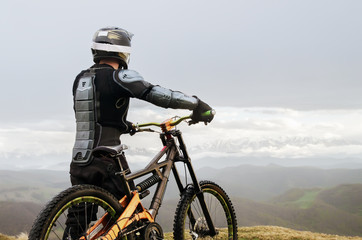 Fototapeta premium The rider in the full-face helmet and full protective equipment on the mtb bike stands on a rock against the background of a ridge and low clouds