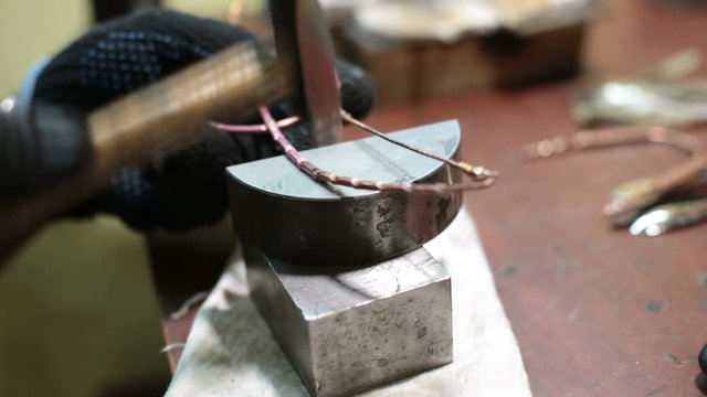 jeweler beats product with hammer on stand in workshop indoors. Close shooting of hands master working in black protective gloves beating thin wire with holes copper thing, upper part in form of