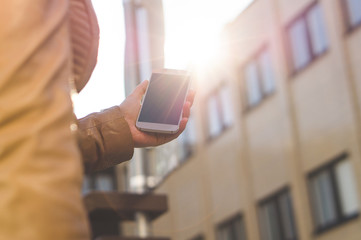 Young woman walking and carrying smartphone in sunshine. Close up of girl holding mobile phone in hand. Morning or evening walk in the city. Urban and modern lifestyle.