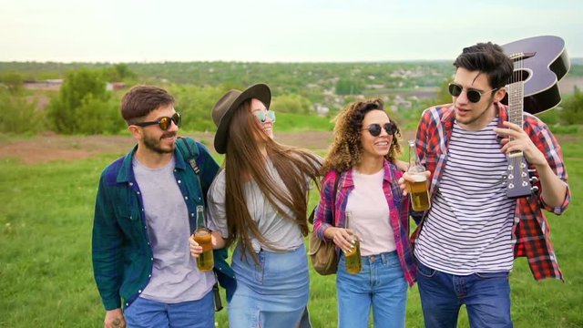 Group of happy hipsters walks forward int the park with great landscape. They very carefree, carrying bottles of beer and guitar