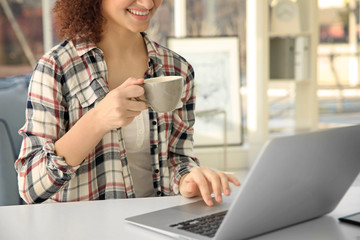 Beautiful young woman with cup of coffee sitting at table and using laptop