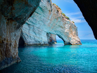 Fototapeta premium Błękitne skały jaskiniowe wysklepiają łuki Błękitnych jaskiń od zwiedzającej łodzi z turystami w błękitnej wodzie Morza Jońskiego wewnątrz jaskini, Wyspa Zakynthos, Grecja wakacje urlopowa wycieczka. Wycieczka z portu Agios Nikolaos