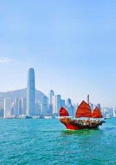 Foto auf Acrylglas Hong Kong Blick auf die Skyline von Hongkong mit einem roten chinesischen Segelboot, das an einem sonnigen Tag am Victoria Harbour vorbeifährt.