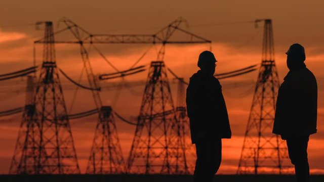 Wide shot of two engineers in hard hat vigorously discussing something at red sunset during an energy substation inspection.