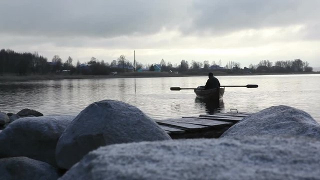 fisherman swims on an old boat to the stony bank at dusk
