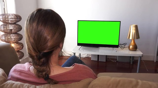 Girl Watching Green Screen Television In Living Room. Young Woman watching television with green screen, shot behind models shoulders