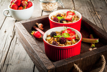 Healthy breakfast. Oatmeal granola crumble with rhubarb, fresh strawberries and blackberries, seeds and ice cream in baked bowls, decorated with mint, on a wooden rustic table in old tray, copy space