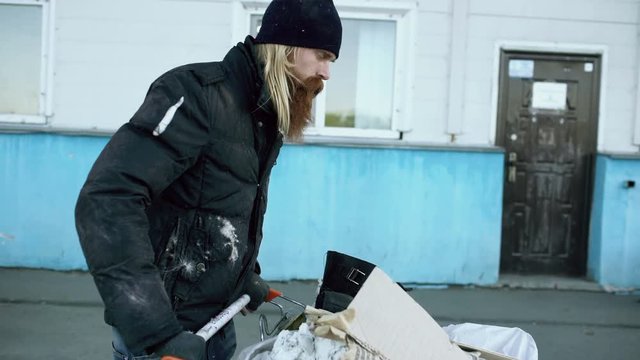 Dolly shot of Bearded homeless man walking down the street with shooping card at cold autumn day